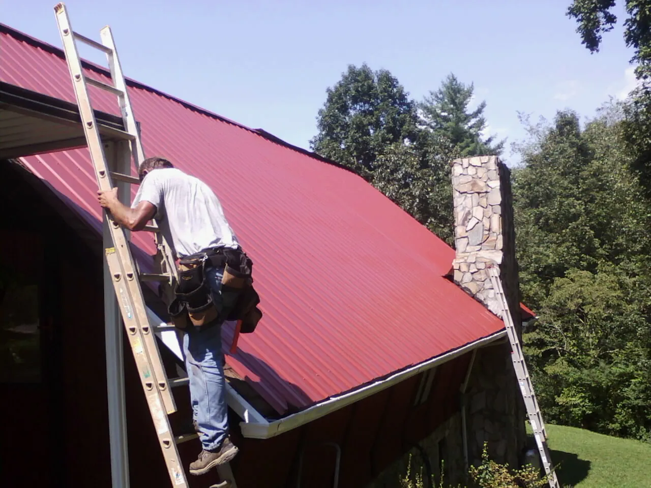 Roofer on a ladder beside a red metal roof and stone chimney — Soffit & Fascia Repair by Roofing Brothers in Dingmans Ferry